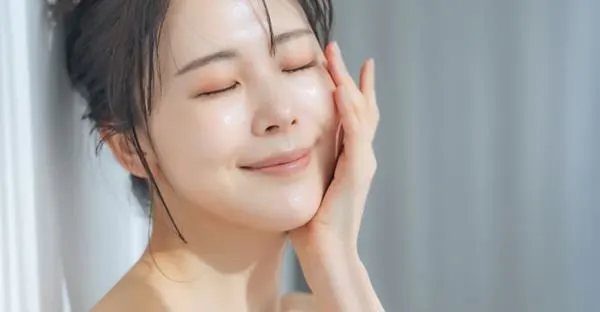 세럼을 바른 후 눈을 감고 미소 짓는 여성의 모습으로, 피부에 은은한 윤광이 돌며 촉촉하고 맑은 피부결이 돋보인다. (A woman smiling softly with her eyes closed after applying serum, her skin glowing with a dewy and radiant finish.)