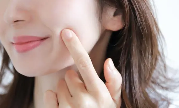 여성의 얼굴을 클로즈업한 장면으로, 손가락으로 볼을 살짝 누르며 피부 탄력을 확인하고 있다. 부드럽고 윤기 있는 피부 질감을 강조한 모습이다. (Close-up of a woman gently pressing her cheek with a finger to check skin elasticity, highlighting smooth and radiant skin texture.)